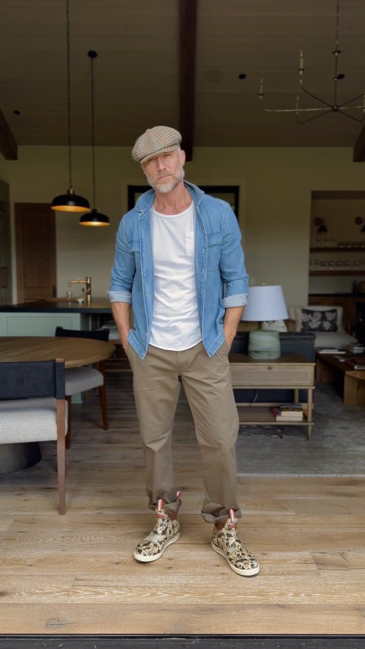 Man wearing a denim shirt, white t-shirt, tan pants, and patterned sneakers standing indoors.
