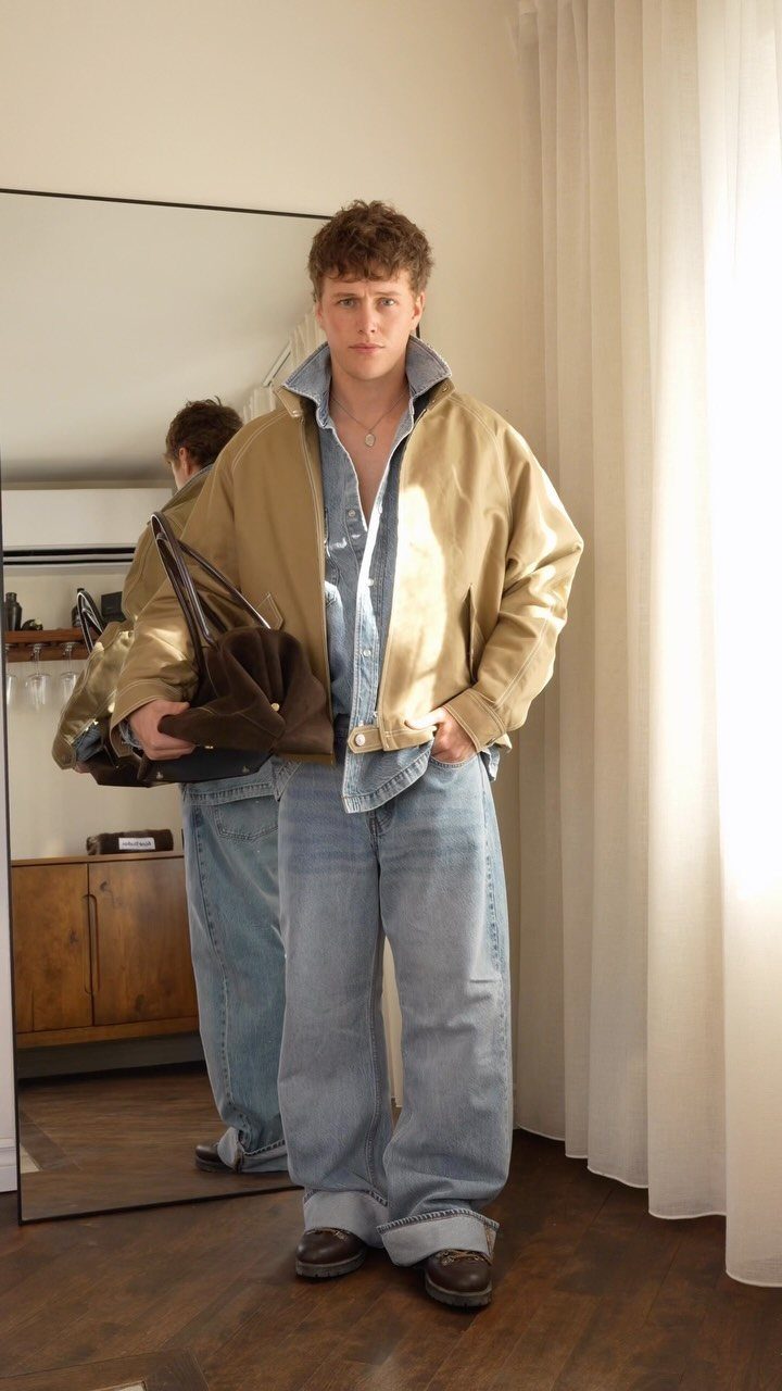 Man wearing a tan bomber jacket over a denim shirt and baggy blue jeans.