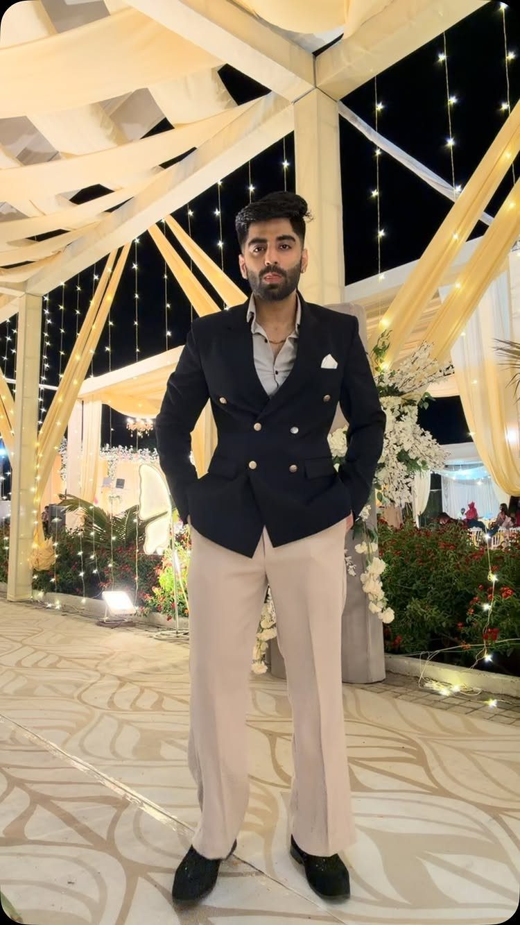Man wearing a navy double-breasted blazer and beige trousers standing under decorative canopy lights.