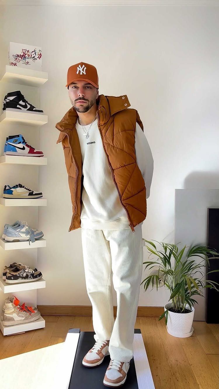 Man wearing a white sweatshirt, white pants, brown puffer vest, and orange cap standing near sneaker shelves.