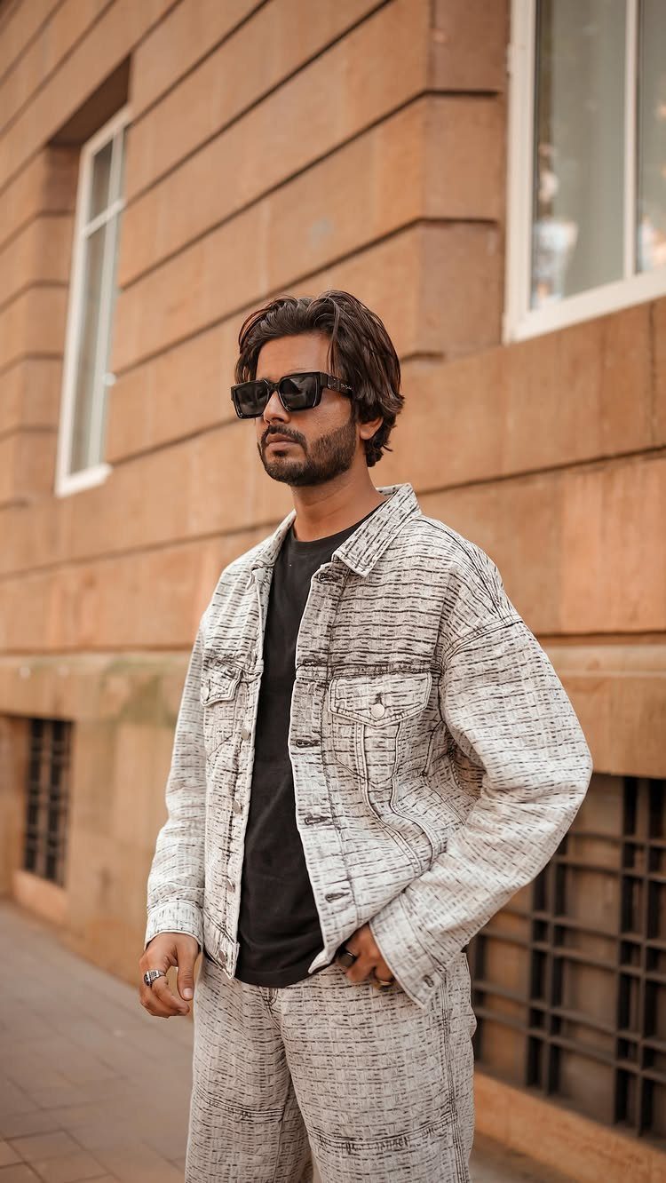 Man in a textured patterned jacket and black t-shirt wearing sunglasses outdoors.