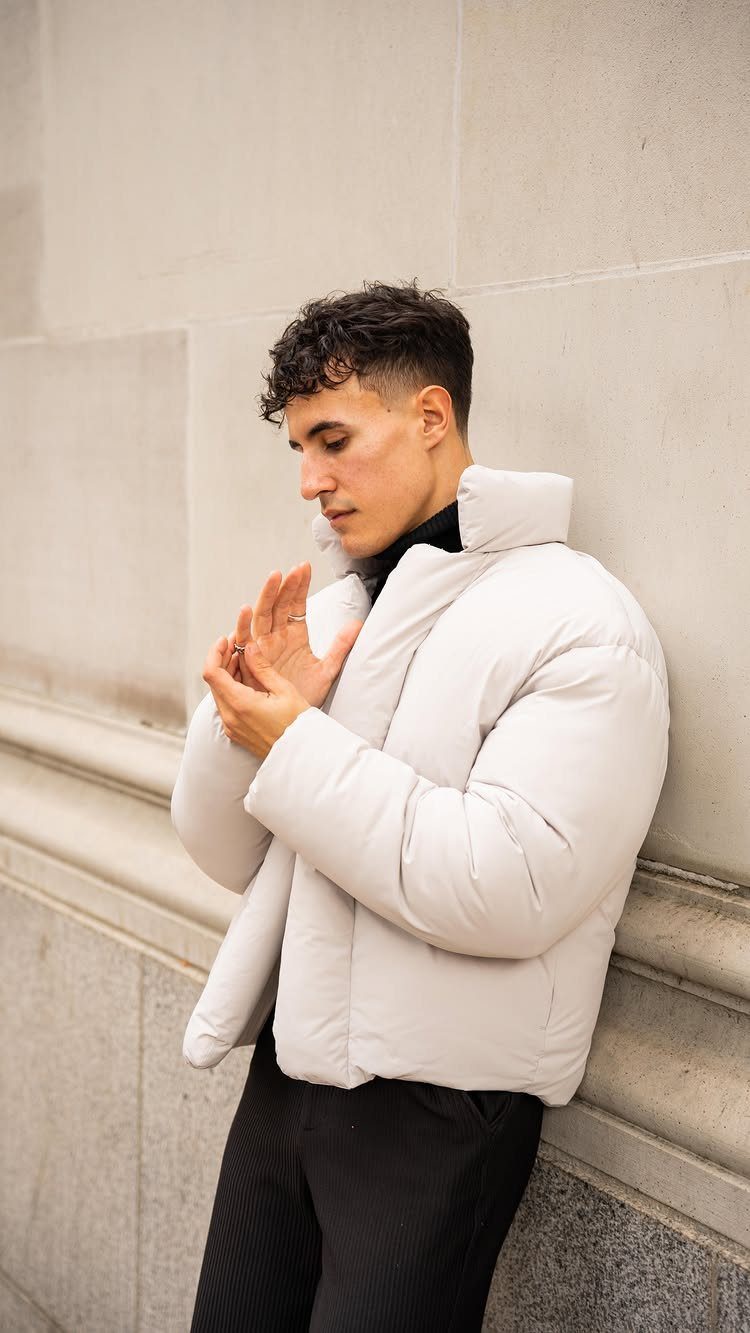 A man in a white puffer jacket and black turtleneck leaning against a stone wall.