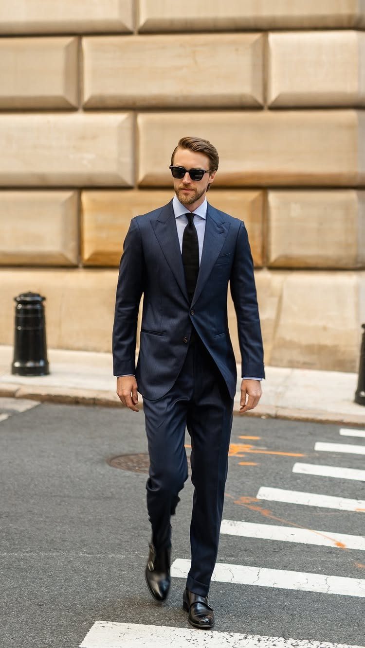 Man in a navy blue suit and sunglasses walking across a city street crosswalk.