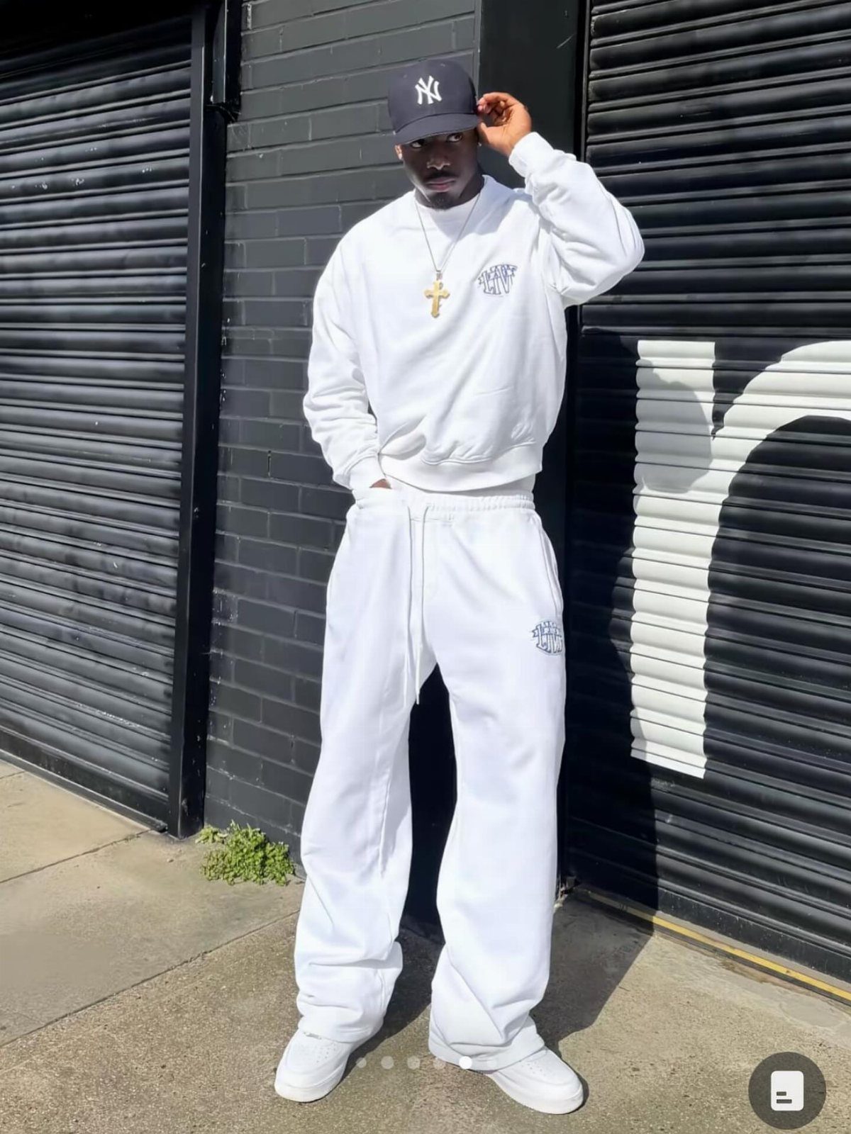 Man in an all-white streetwear outfit posing against black shutters.