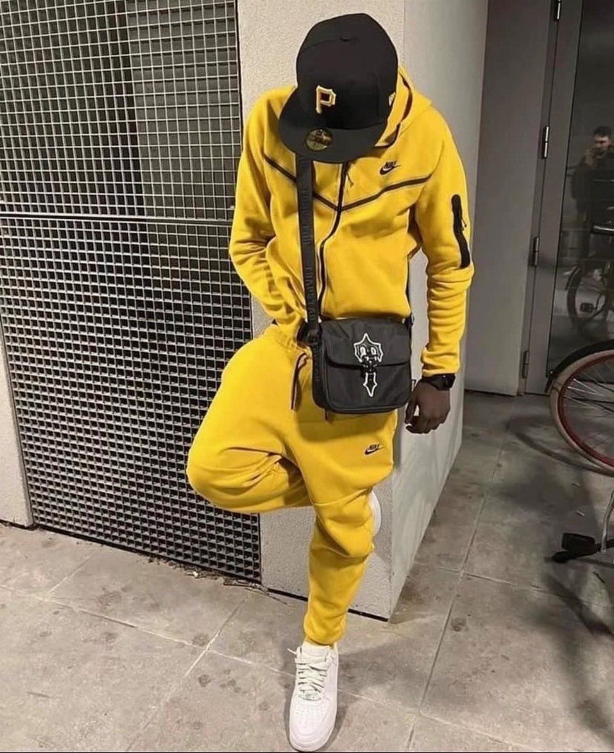 A person in a yellow tracksuit and black baseball cap posing against a metal wall.