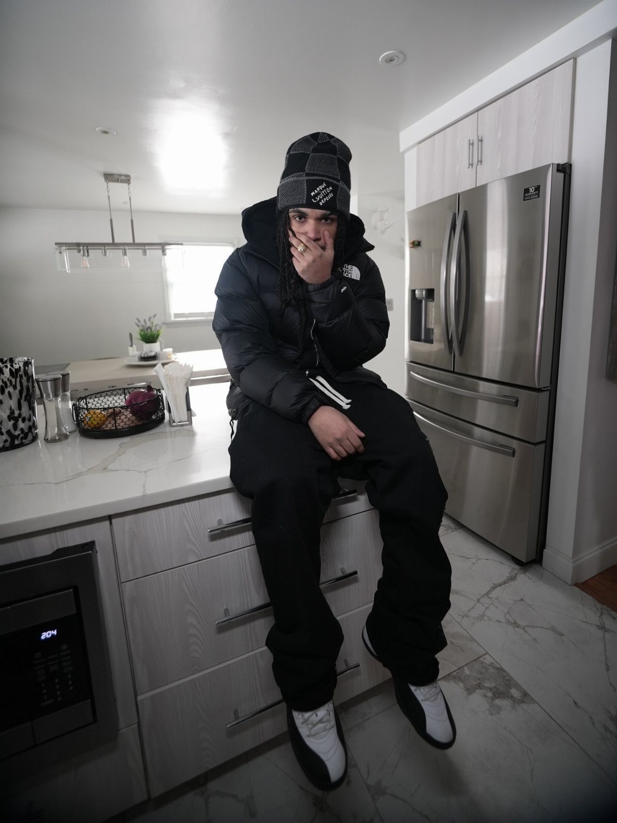 Man in black puffer jacket and patterned beanie sitting on a white kitchen counter.