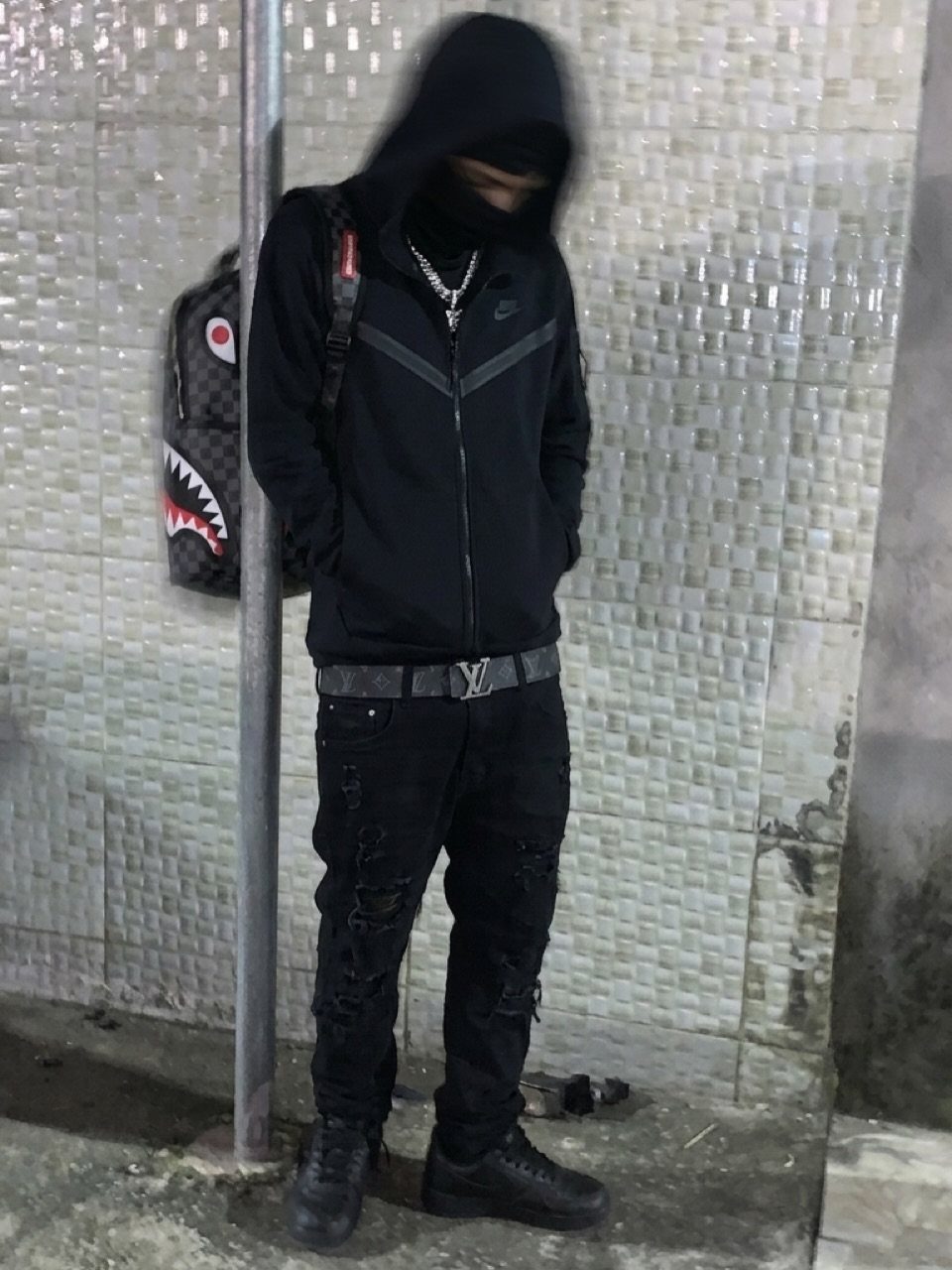 Man in black and grey zip-up hoodie, silver chains, and black distressed pants standing by a tiled wall.