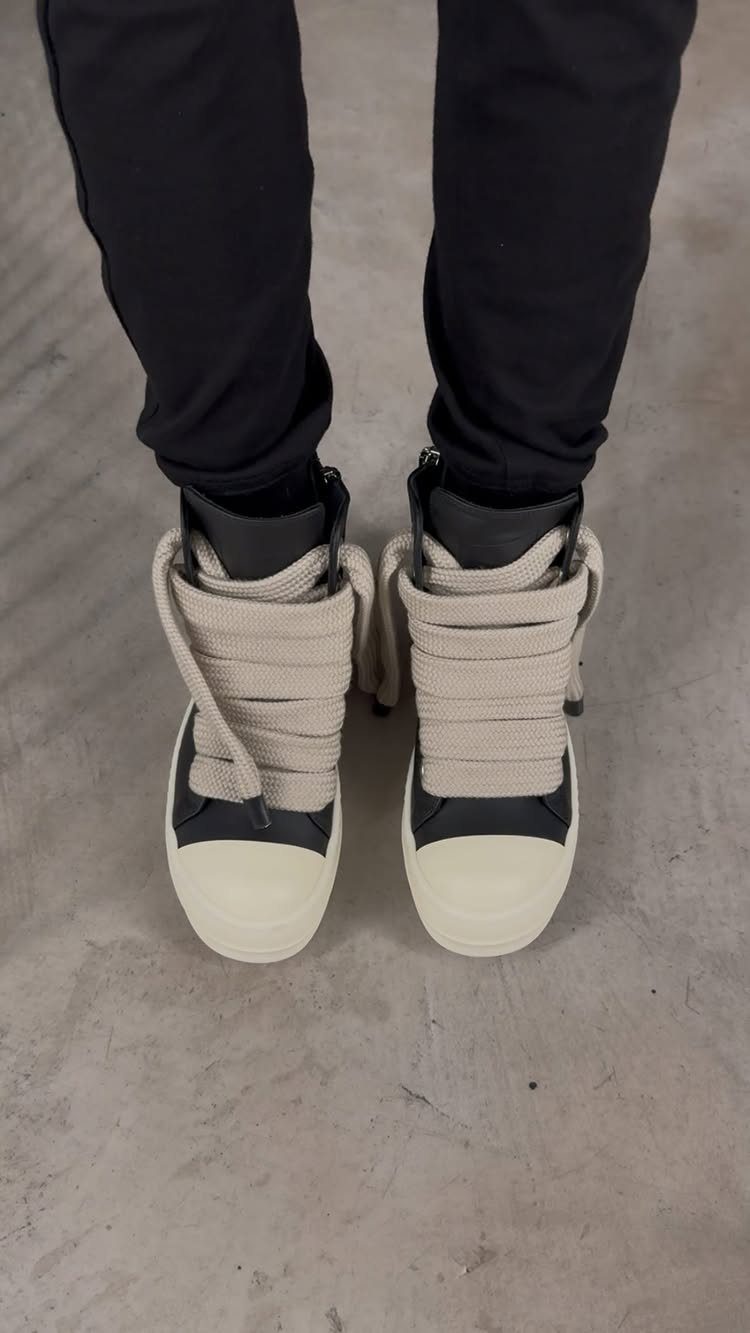 Black and white high-top sneakers with thick cream rope laces worn with black pants.