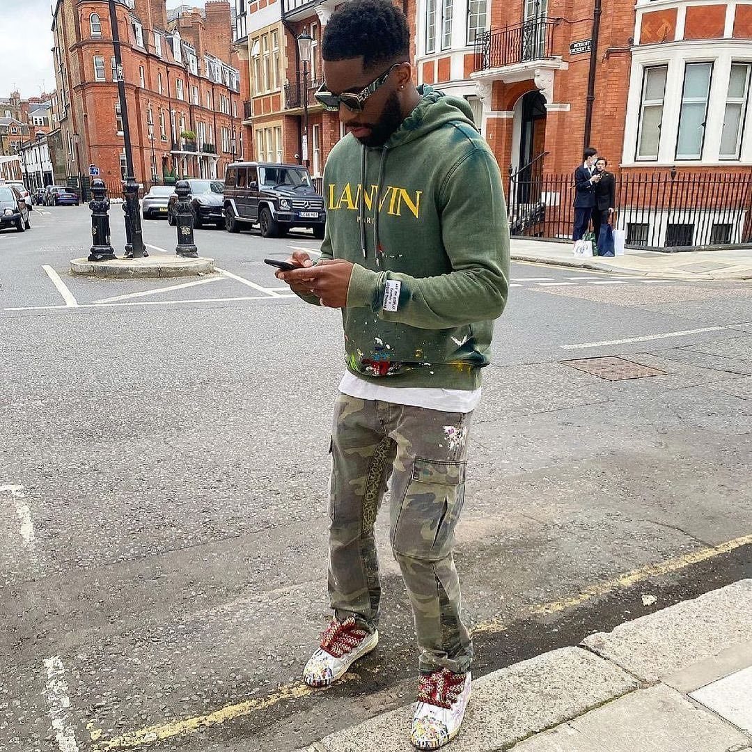Man in olive green hoodie and camouflage cargo pants standing on a city street.
