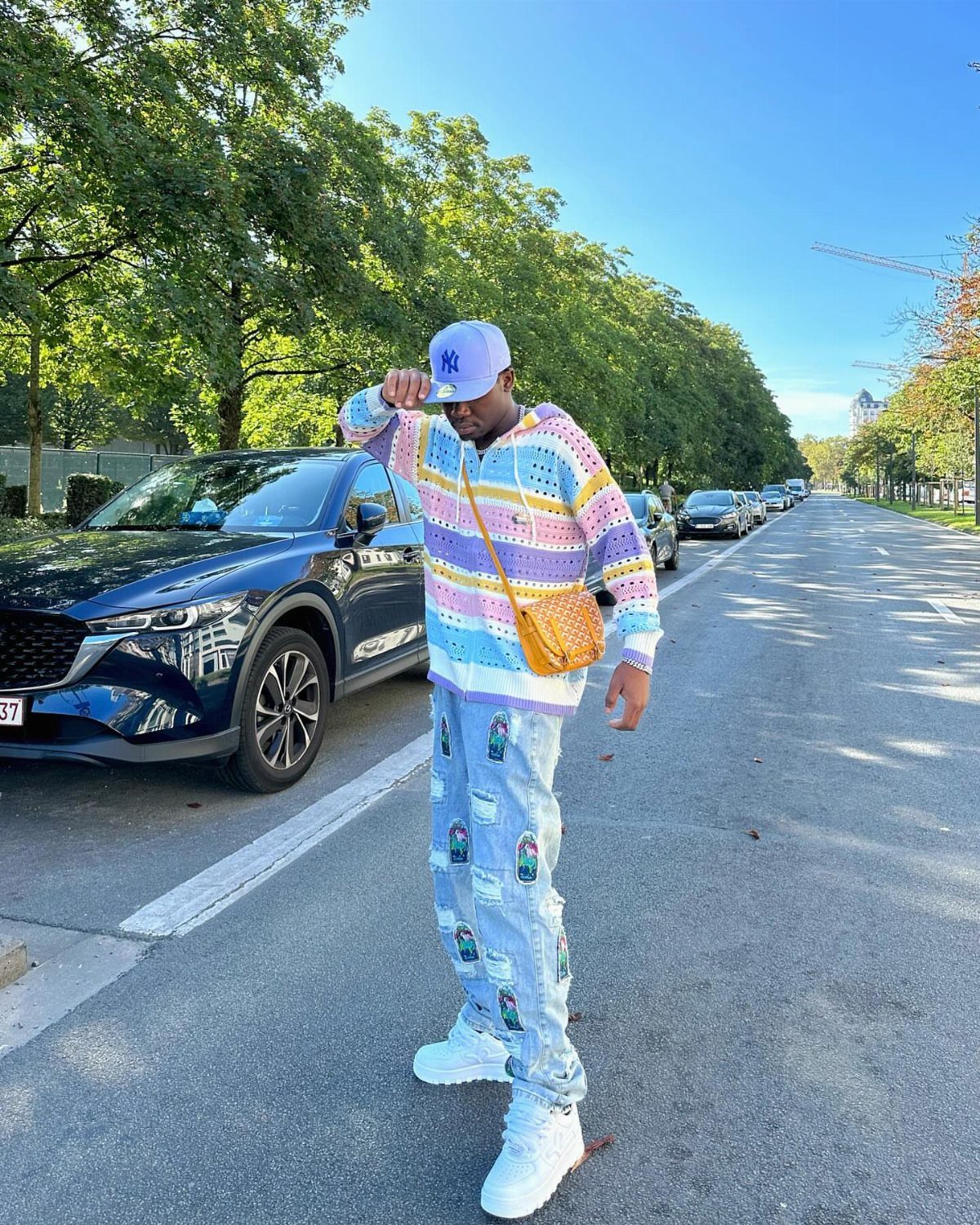 Man in a colorful crochet hoodie, patterned pants, and white sneakers standing on a street.
