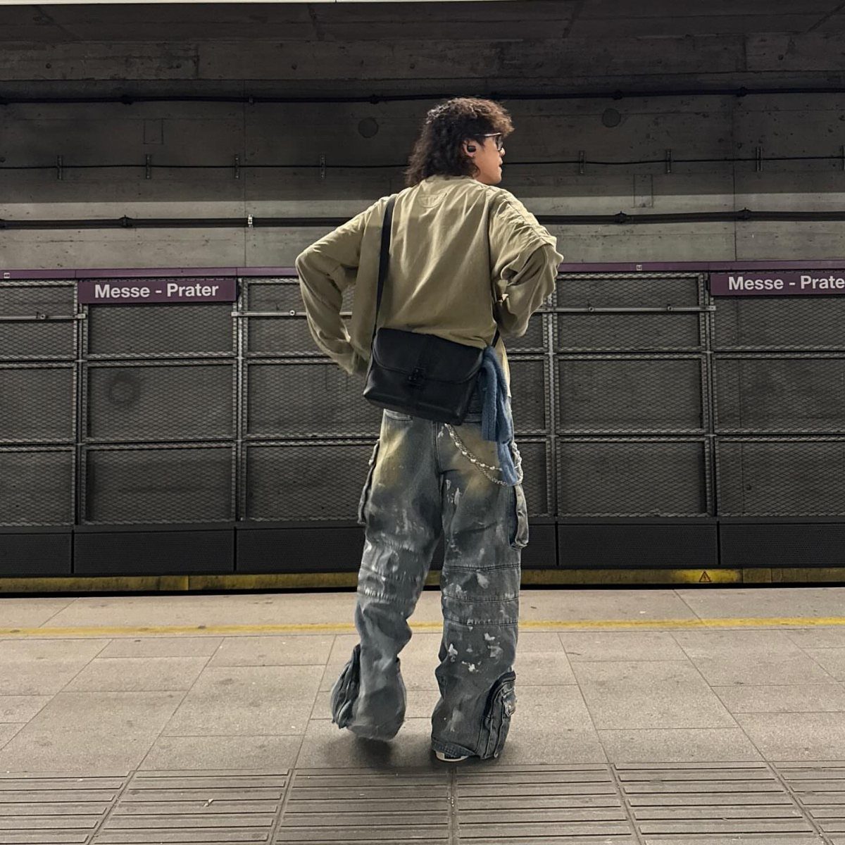 Man in tan shirt and baggy distressed denim cargo jeans standing in a subway station.