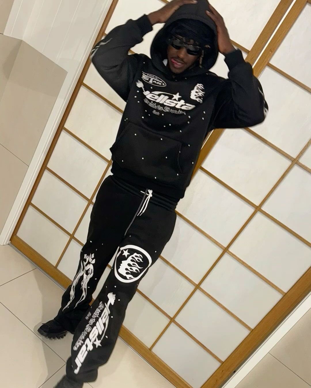 Man in a black graphic hoodie and matching joggers posing against a shoji screen.