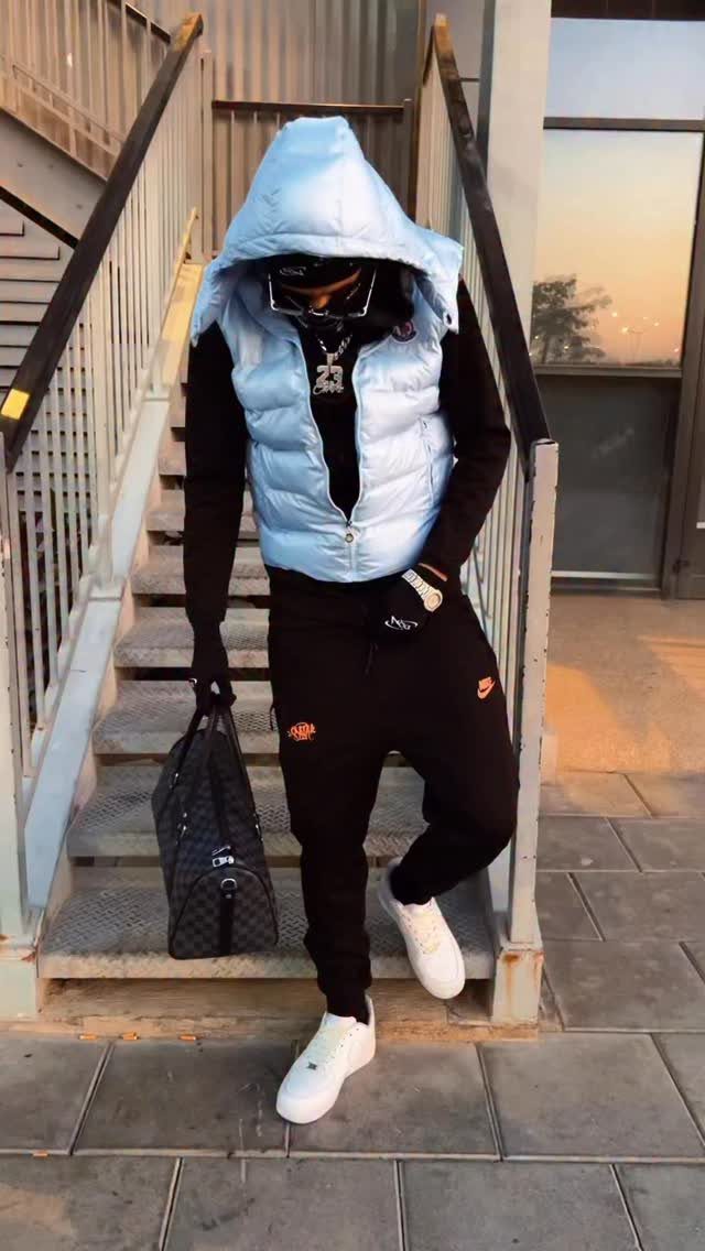 Man in light blue puffer vest, black joggers, and white sneakers standing on stairs.