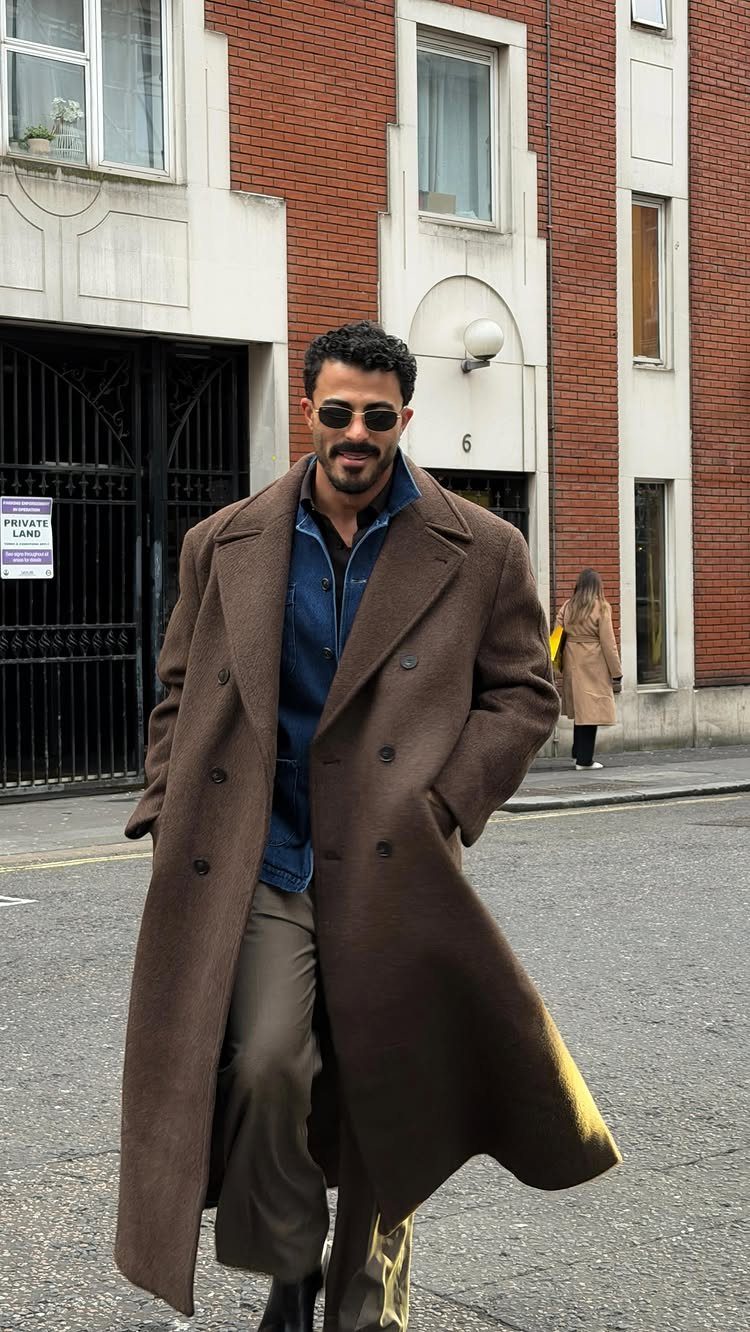 A man wearing a long brown wool overcoat and sunglasses walking on a city street.