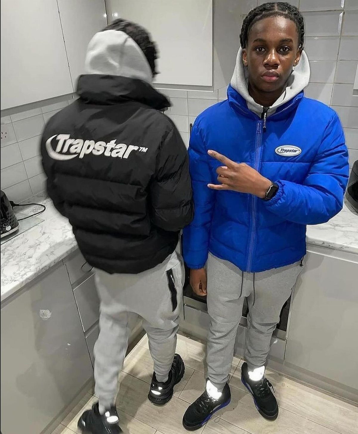 Two men wearing Trapstar brand puffer jackets and grey joggers in a kitchen.
