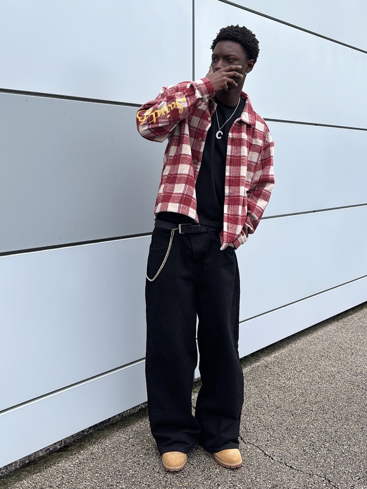 Man wearing a red plaid flannel shirt, black t-shirt, and black trousers against a blue wall.