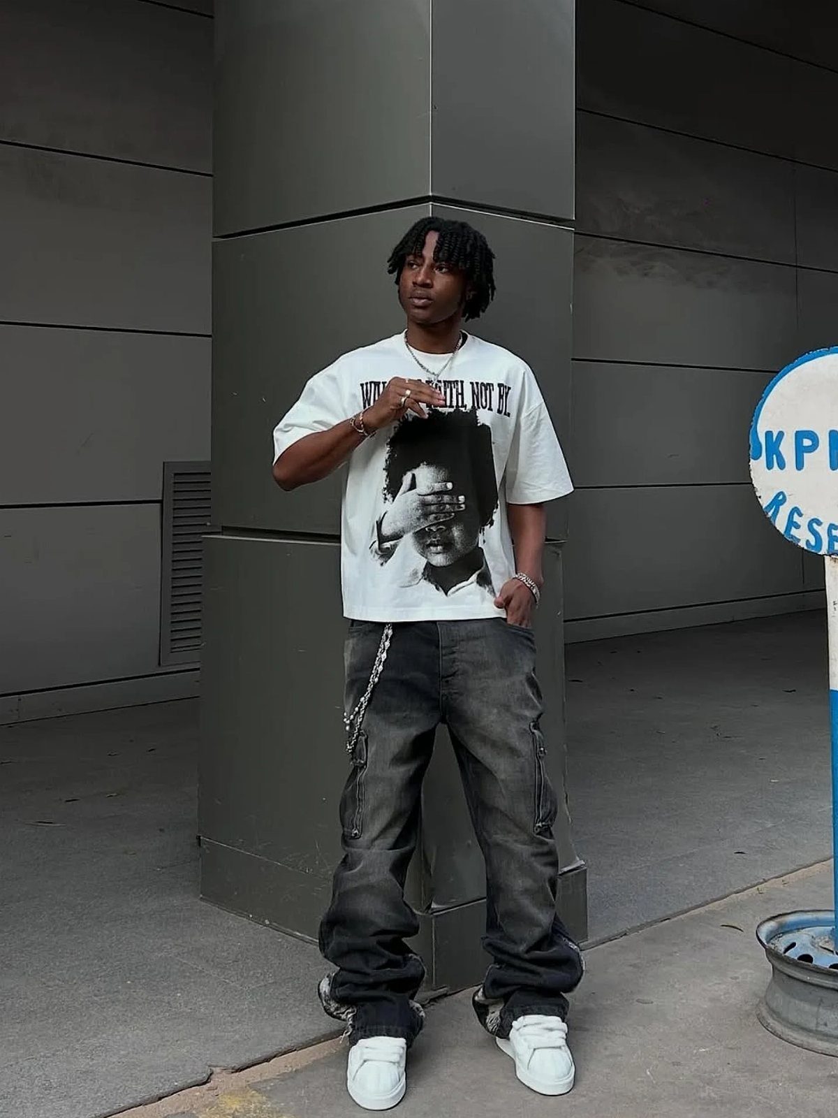 A man posing in a white graphic t-shirt, dark cargo pants, and white sneakers.