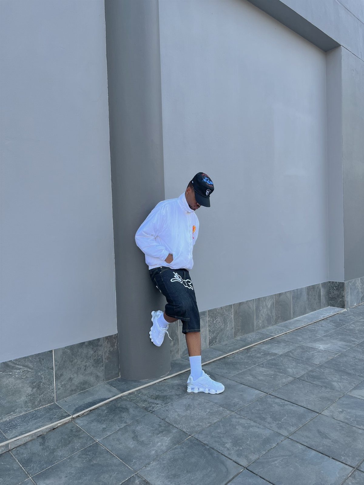 Man in white shirt and black shorts walking in white sneakers against a grey wall.