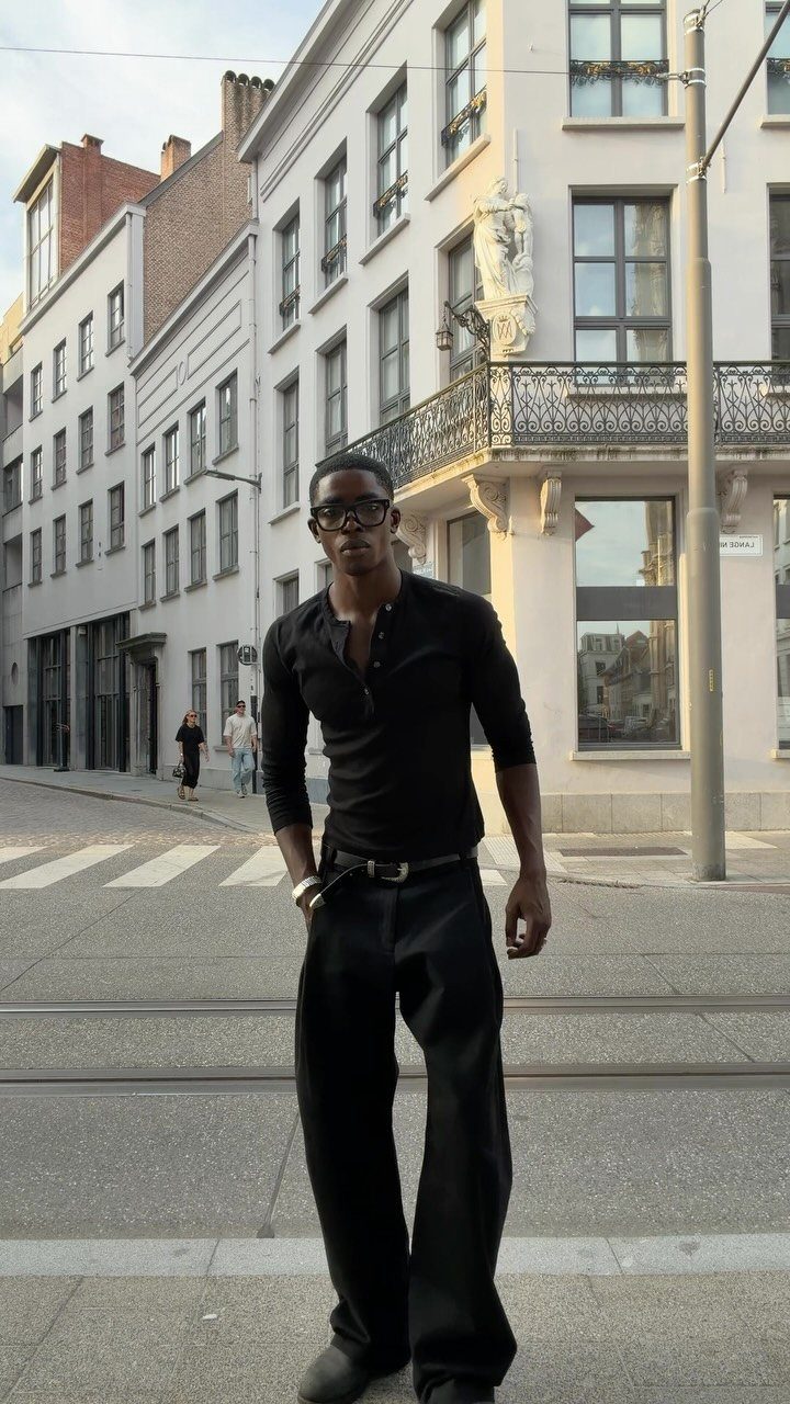 A man in a black henley shirt and black trousers posing on a city street.