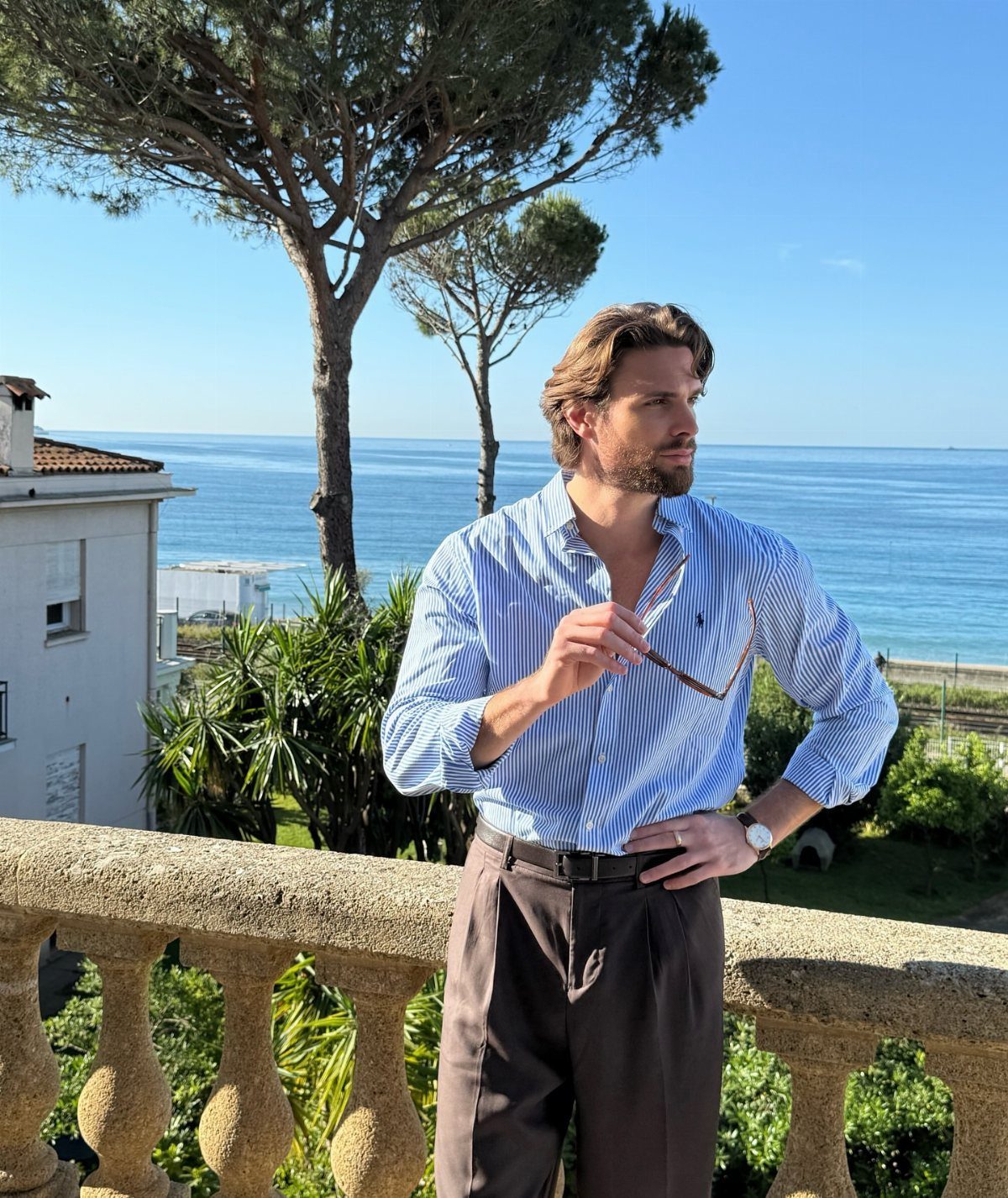 Man in a striped shirt and brown trousers posing on a balcony by the sea.