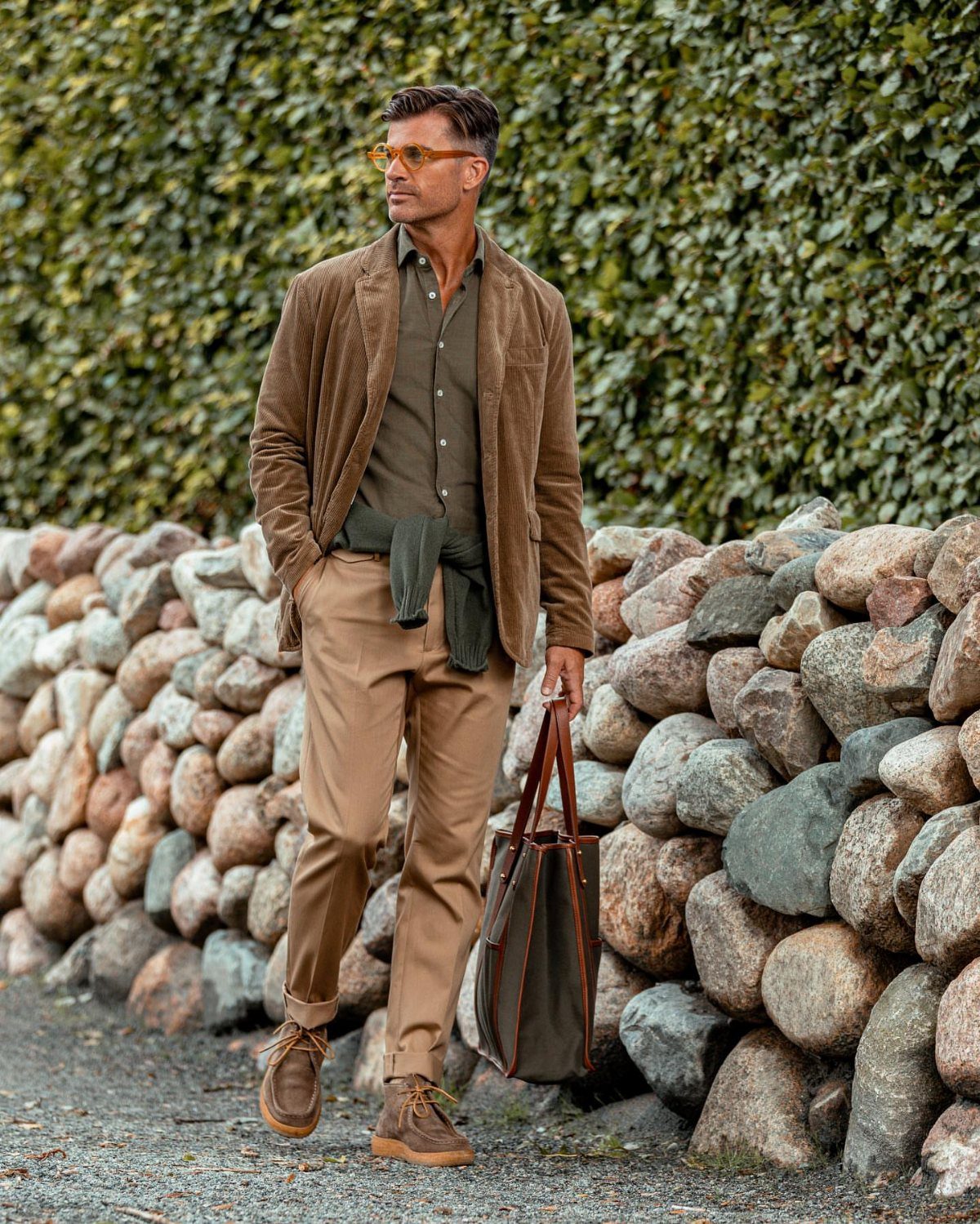 Man wearing a brown corduroy blazer, olive shirt, and tan trousers standing by a stone wall.