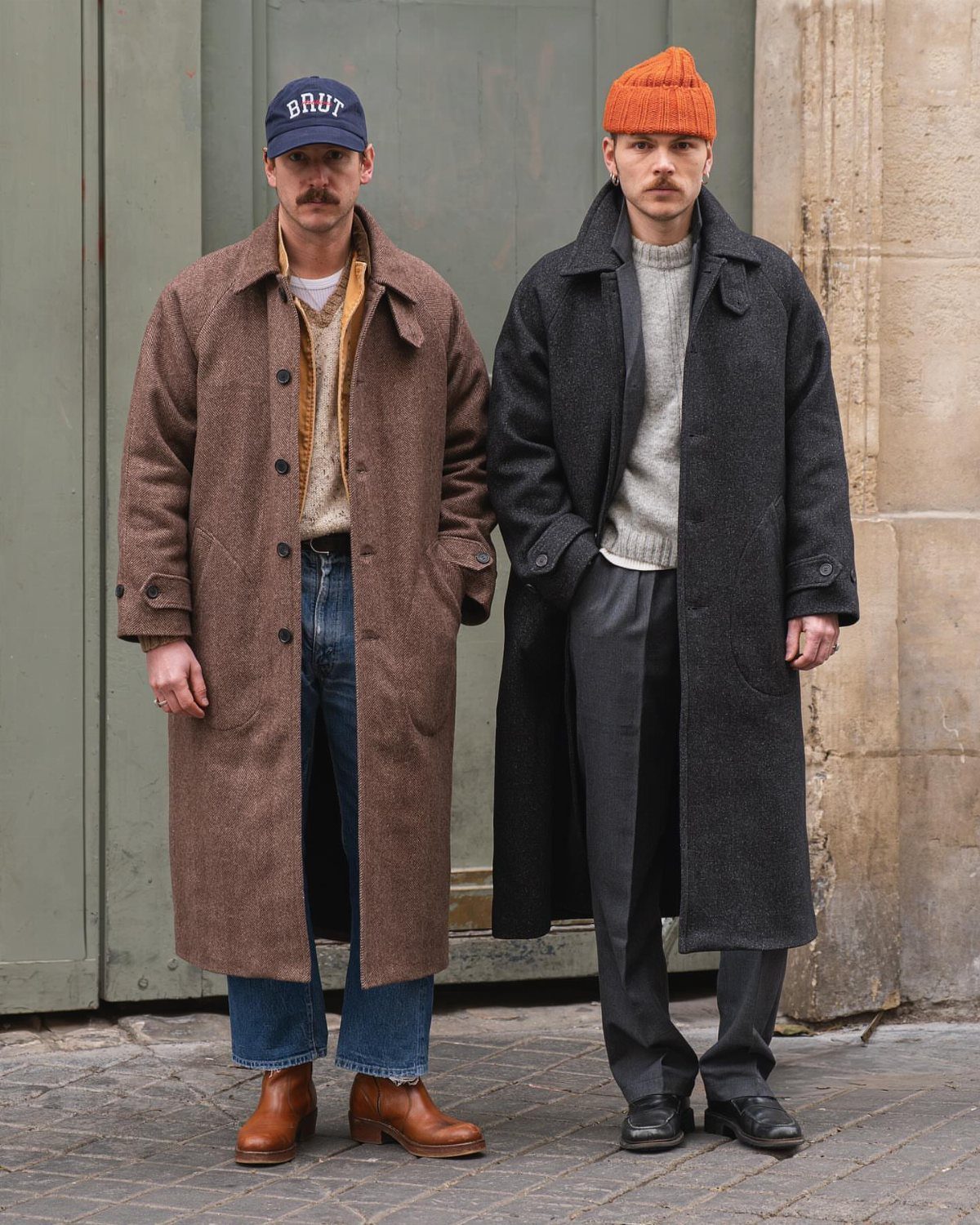 Two men standing side by side wearing long, stylish heavy overcoats and winter accessories.