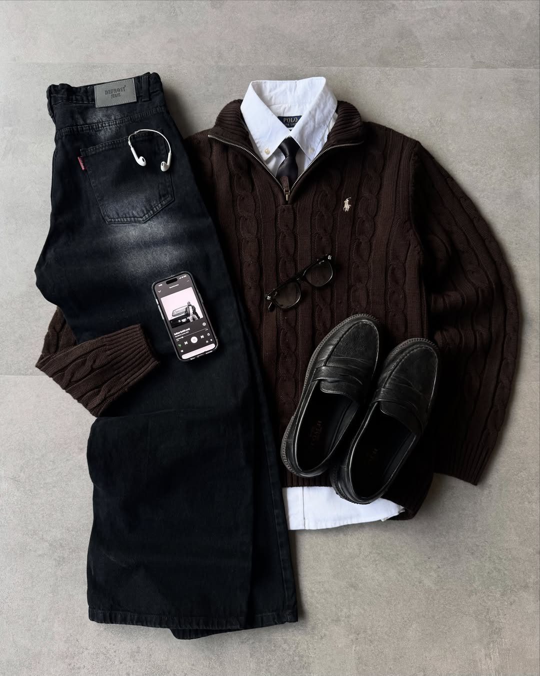 Brown cable-knit sweater, white shirt, tie, dark jeans, and black loafers arranged in a flat lay.