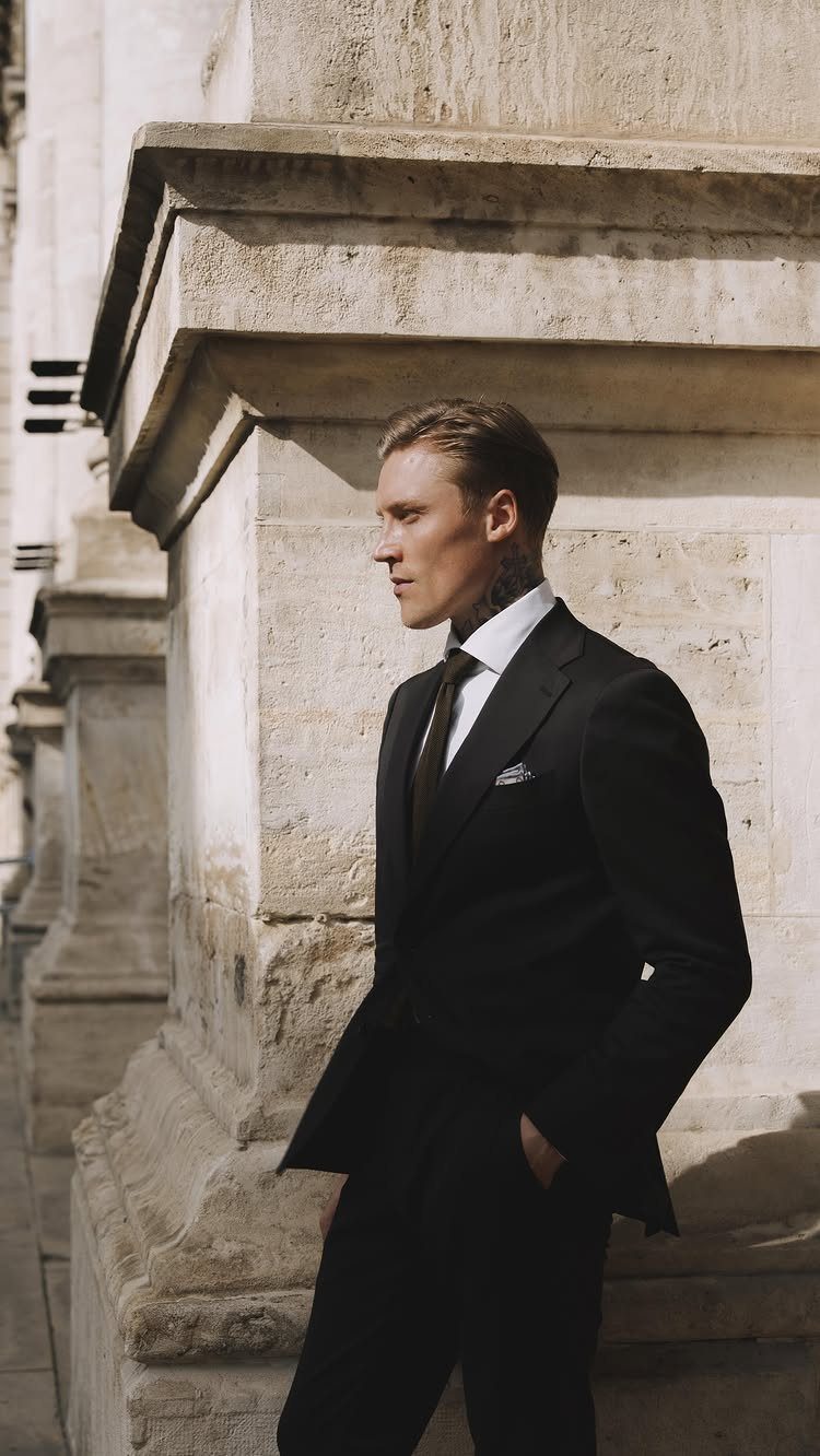 A man wearing a black tailored suit and tie standing by a stone pillar.