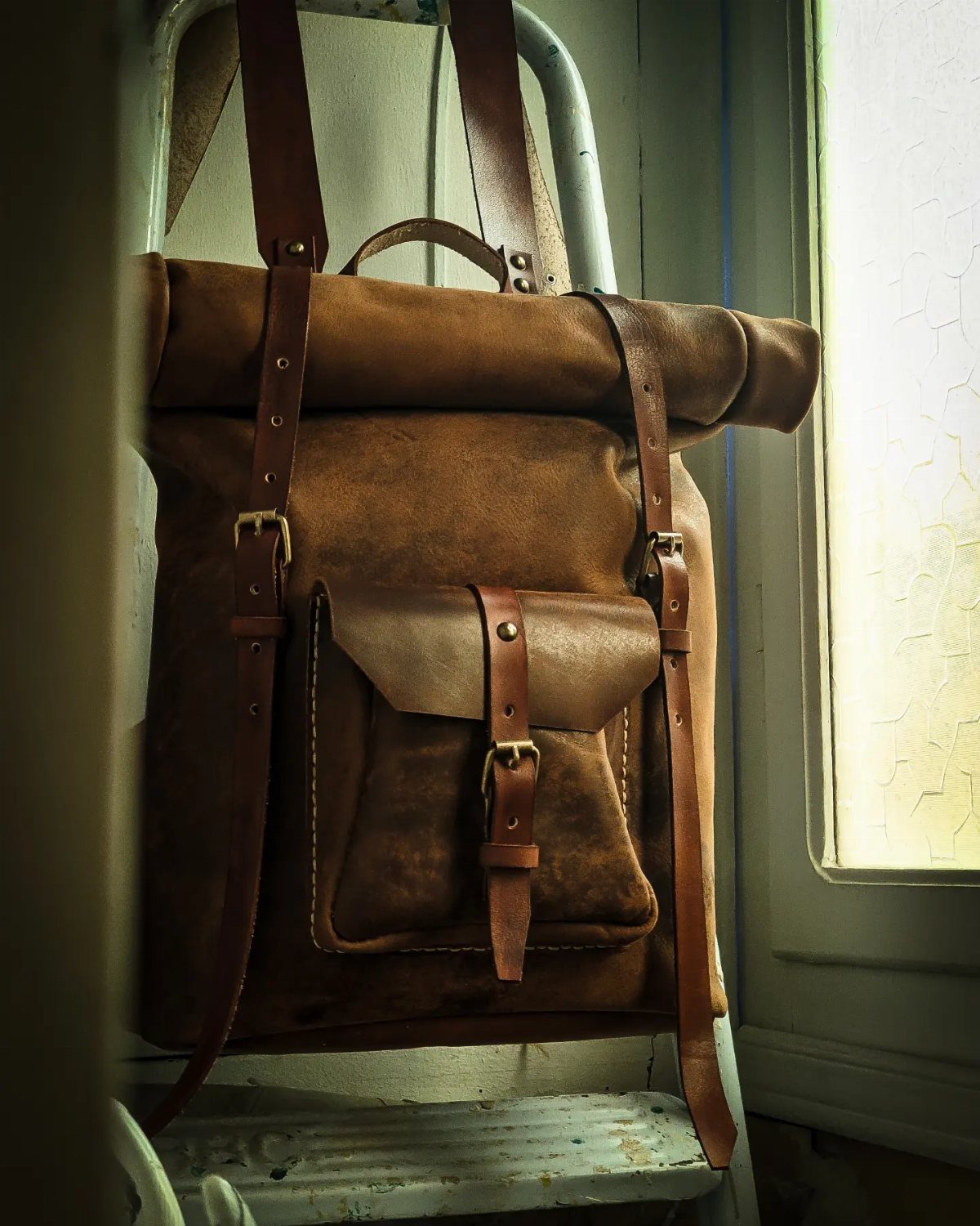A rustic brown leather roll-top backpack hanging on a white metal chair by a window.
