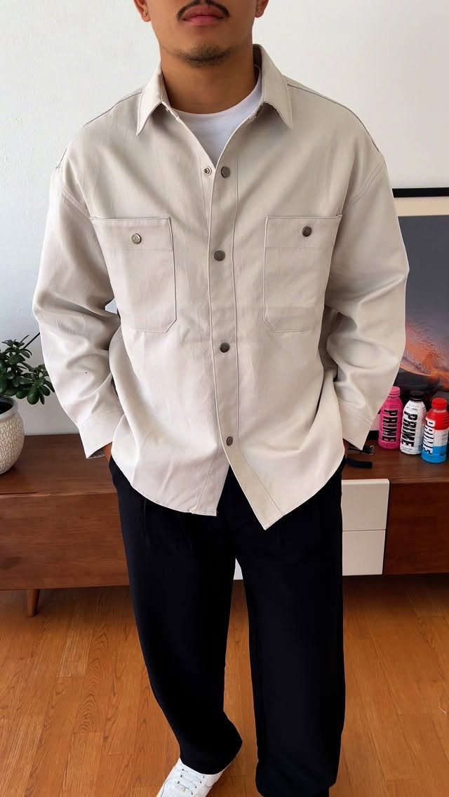 A man wearing a beige long-sleeved overshirt with large chest pockets and black trousers.