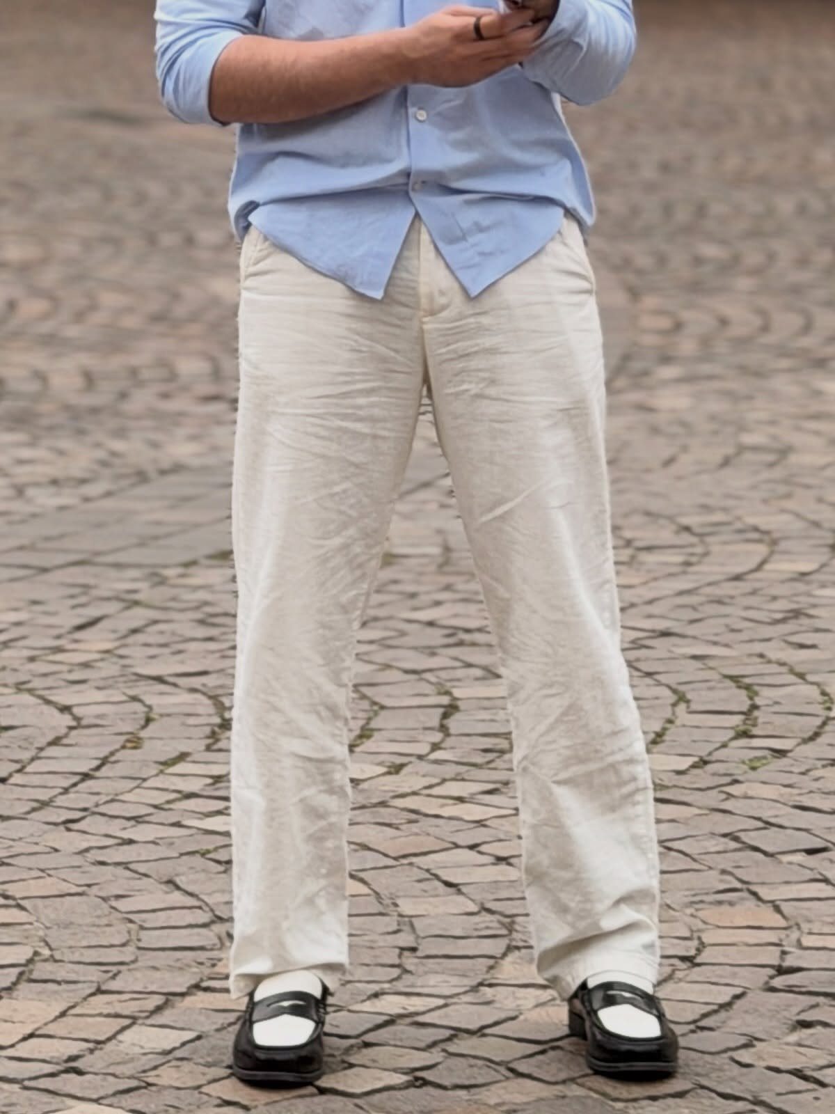 Man wearing a light blue shirt, cream linen pants, and black and white loafers on cobblestones.