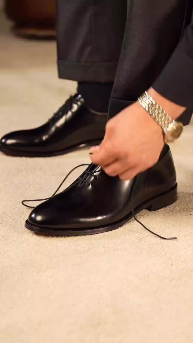 A person tying the laces of polished black leather derby shoes while wearing dark trousers.