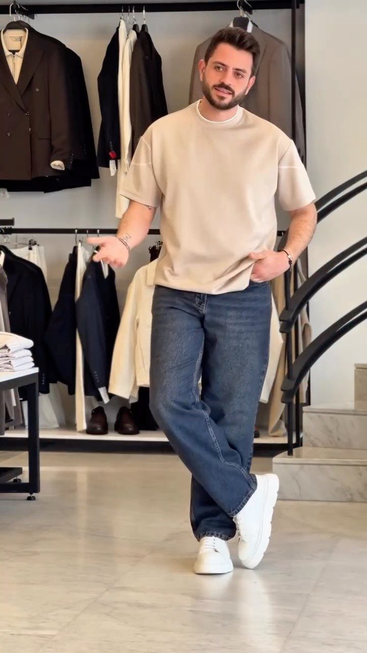 A man posing in a beige oversized t-shirt and blue jeans inside a clothing boutique.