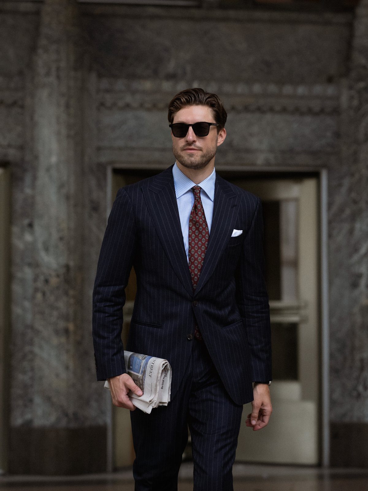 A man in a navy pinstriped suit, light blue shirt, and burgundy tie walking outdoors.