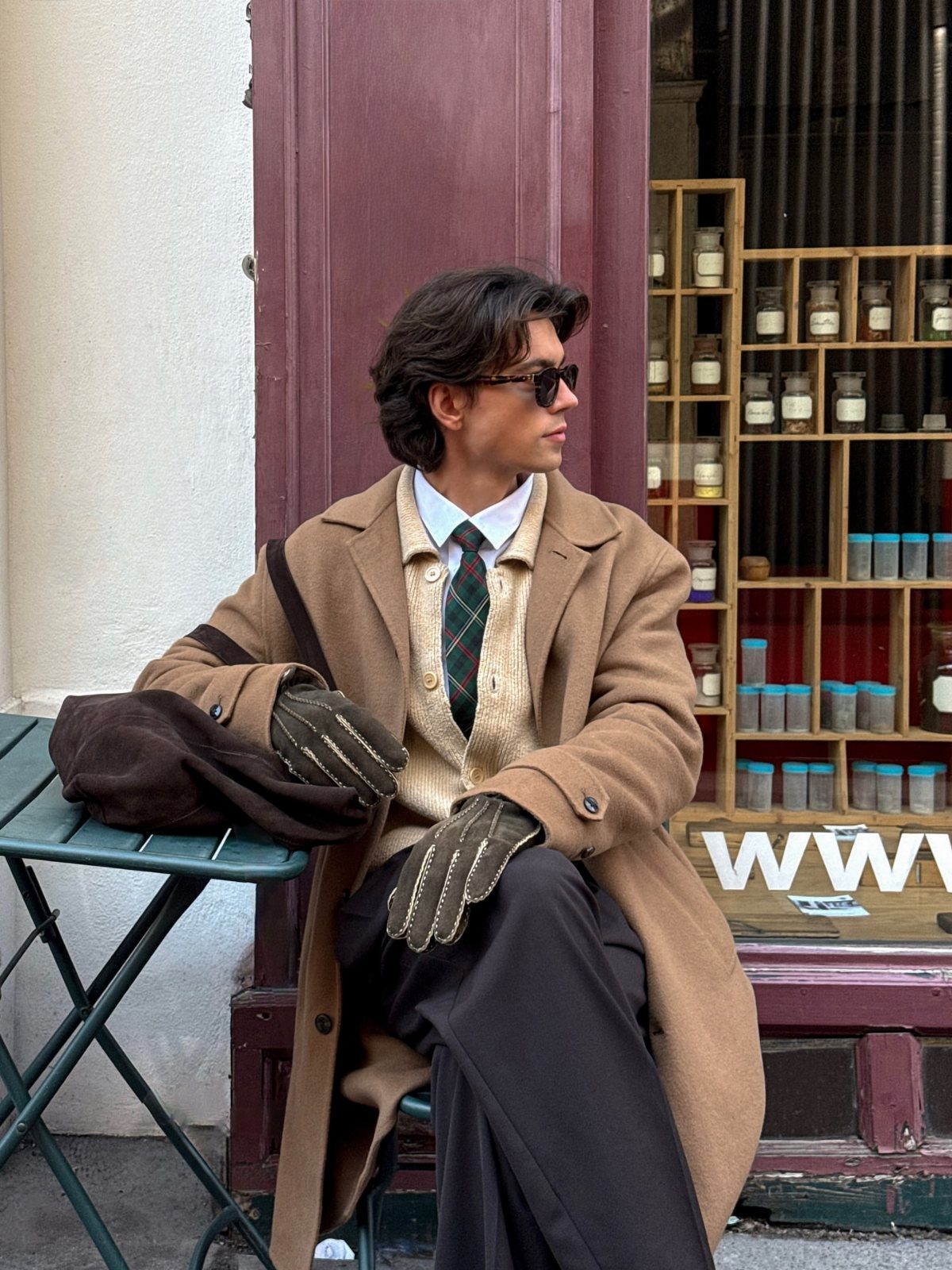 Man in a tan wool coat, cream sweater, and plaid tie sitting by a shop.
