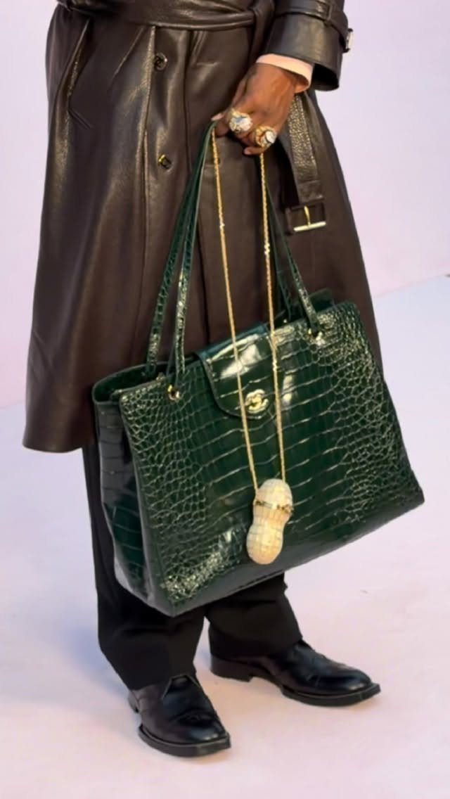 A person holding a large green crocodile-textured handbag while wearing chunky gold rings and a statement pendant necklace.
