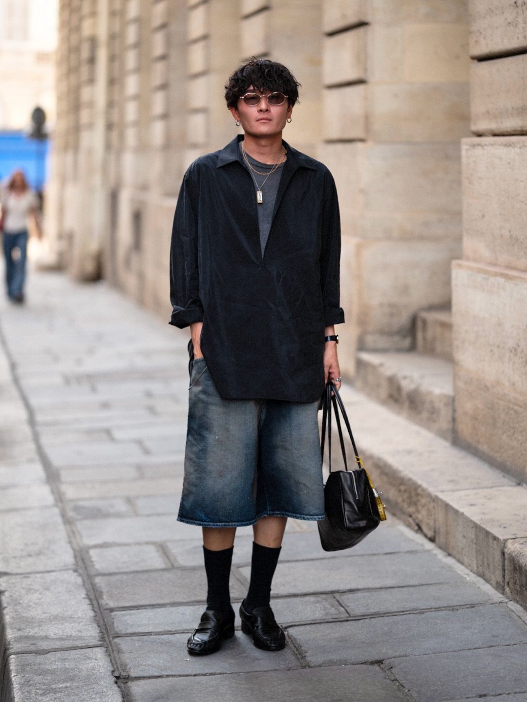 A person posing in an oversized black shirt, denim shorts, and black loafers on a city street.