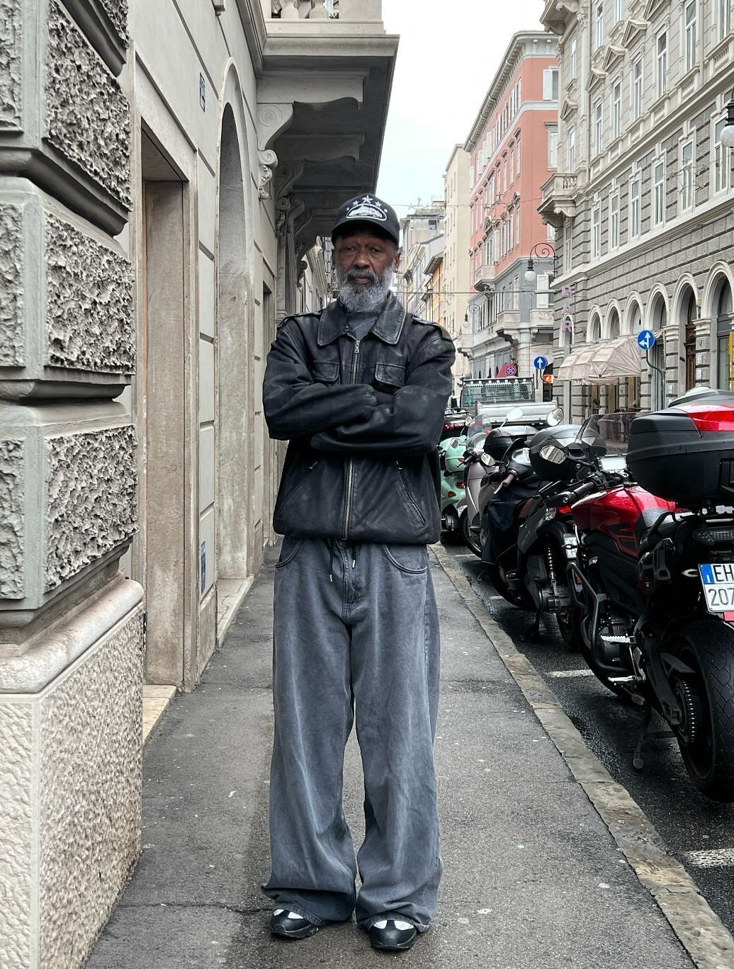 An older man with a grey beard wearing a leather jacket and baggy denim jeans standing on a city street.