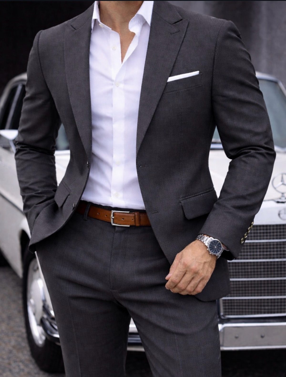 Man in a charcoal grey fitted suit and white shirt standing by a vintage car.