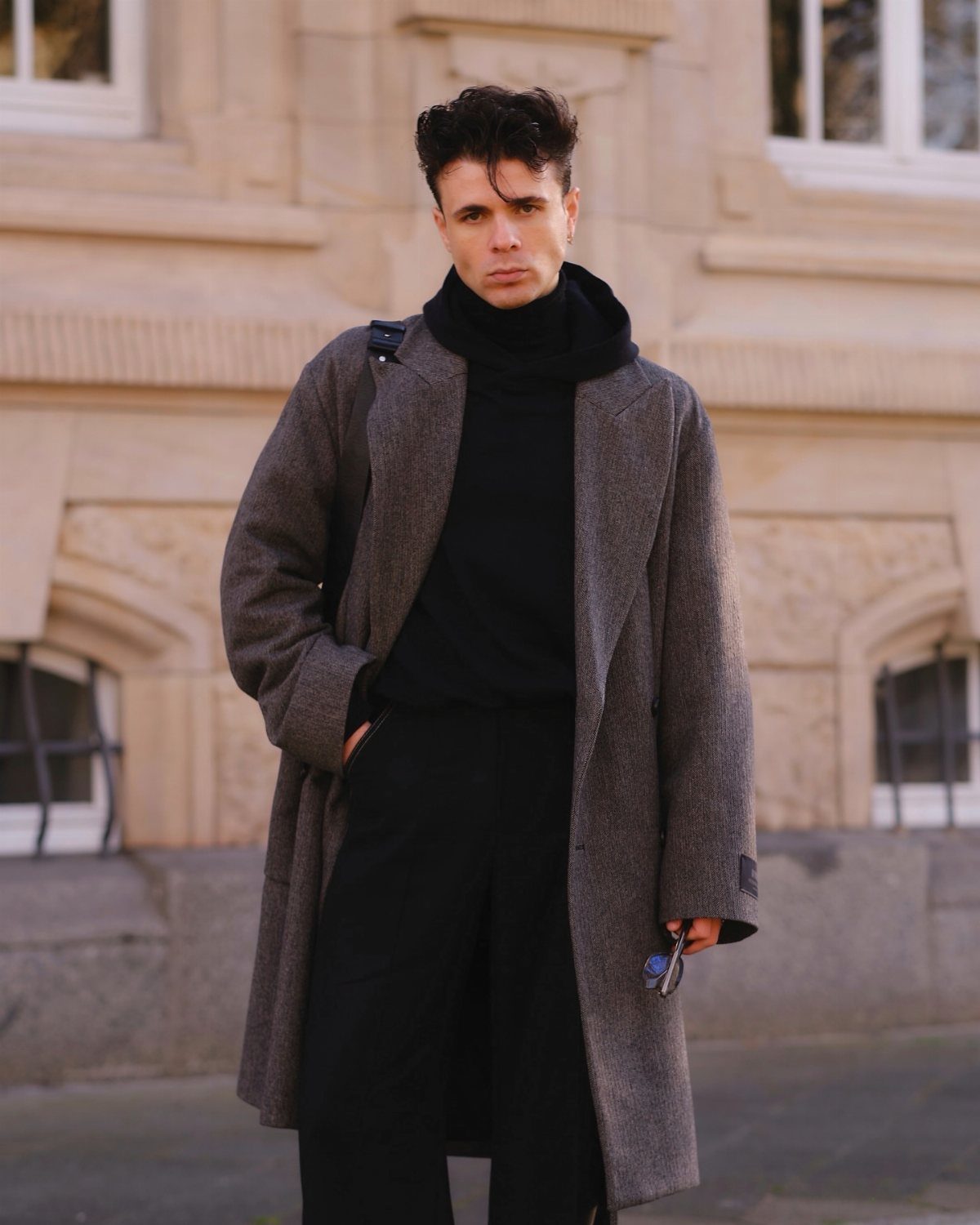 A man wearing a long grey wool coat and black turtleneck standing in front of a building.