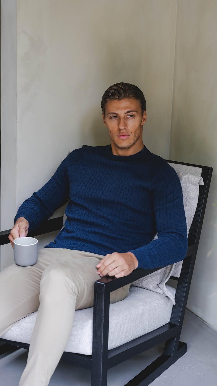 A man in a navy blue sweater and beige trousers sitting in a chair holding a mug.