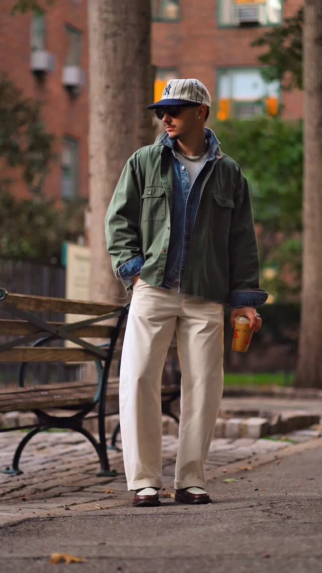 Man wearing an olive green utility jacket, denim shirt, and white wide-leg trousers standing in a park.