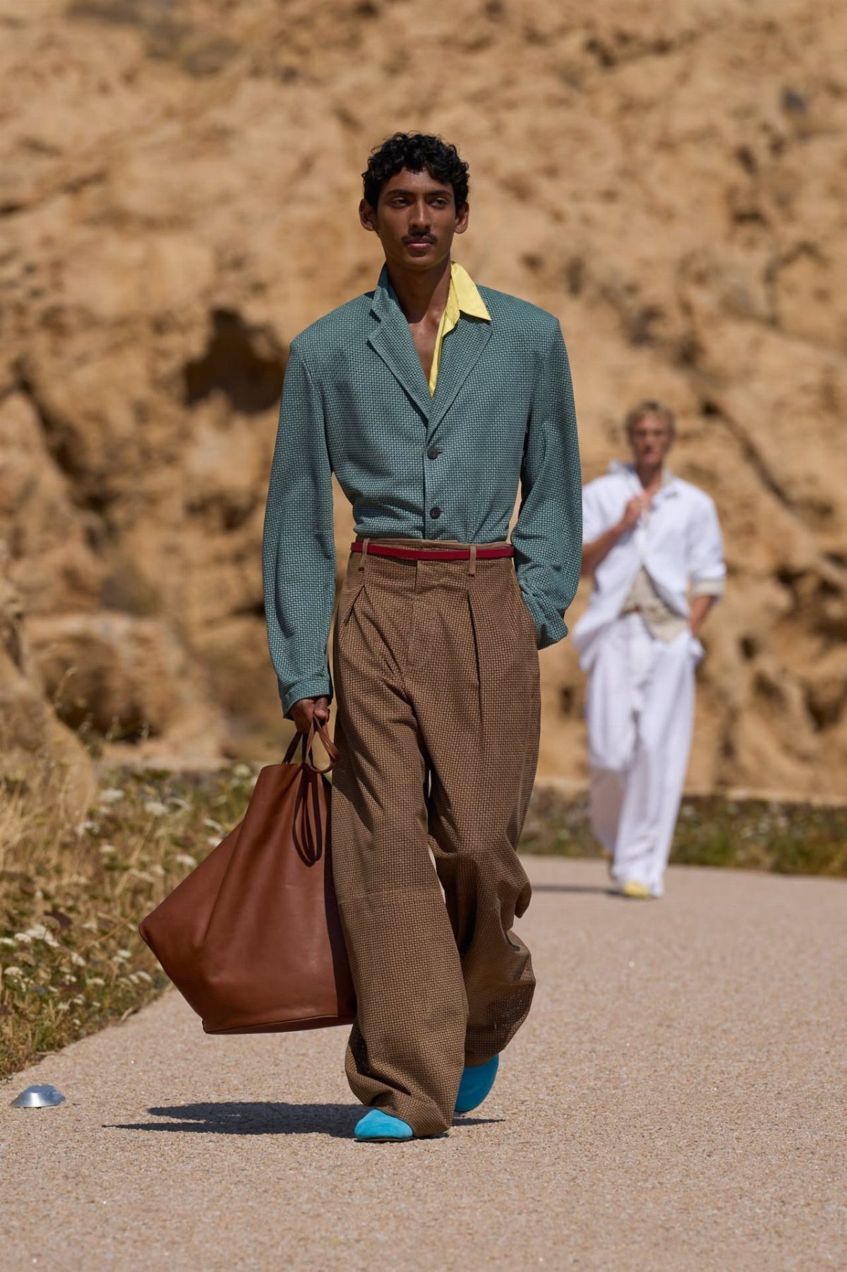Man walking in a teal shirt, yellow collar, brown trousers, and blue shoes outdoors.