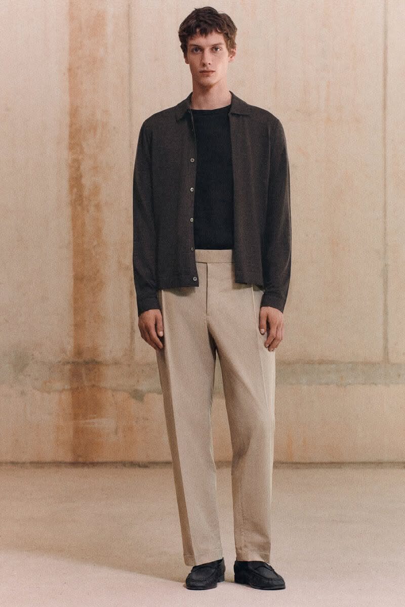 A man wearing a dark shirt, black t-shirt, and beige trousers standing against a textured wall.