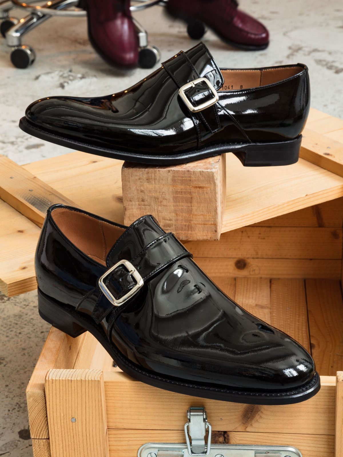 Two black patent leather monk strap shoes with silver buckles resting on wooden crates.
