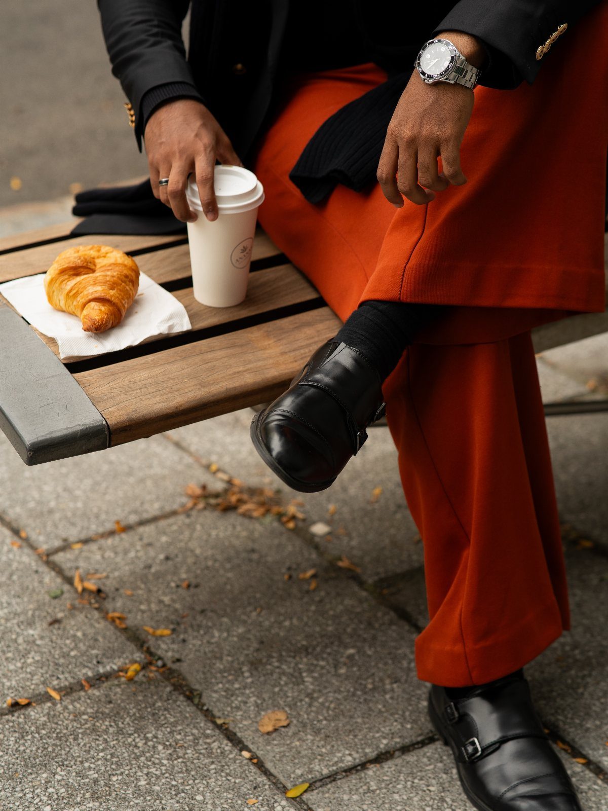 Person wearing bold orange trousers and black blazer sitting on a bench with coffee and a croissant.