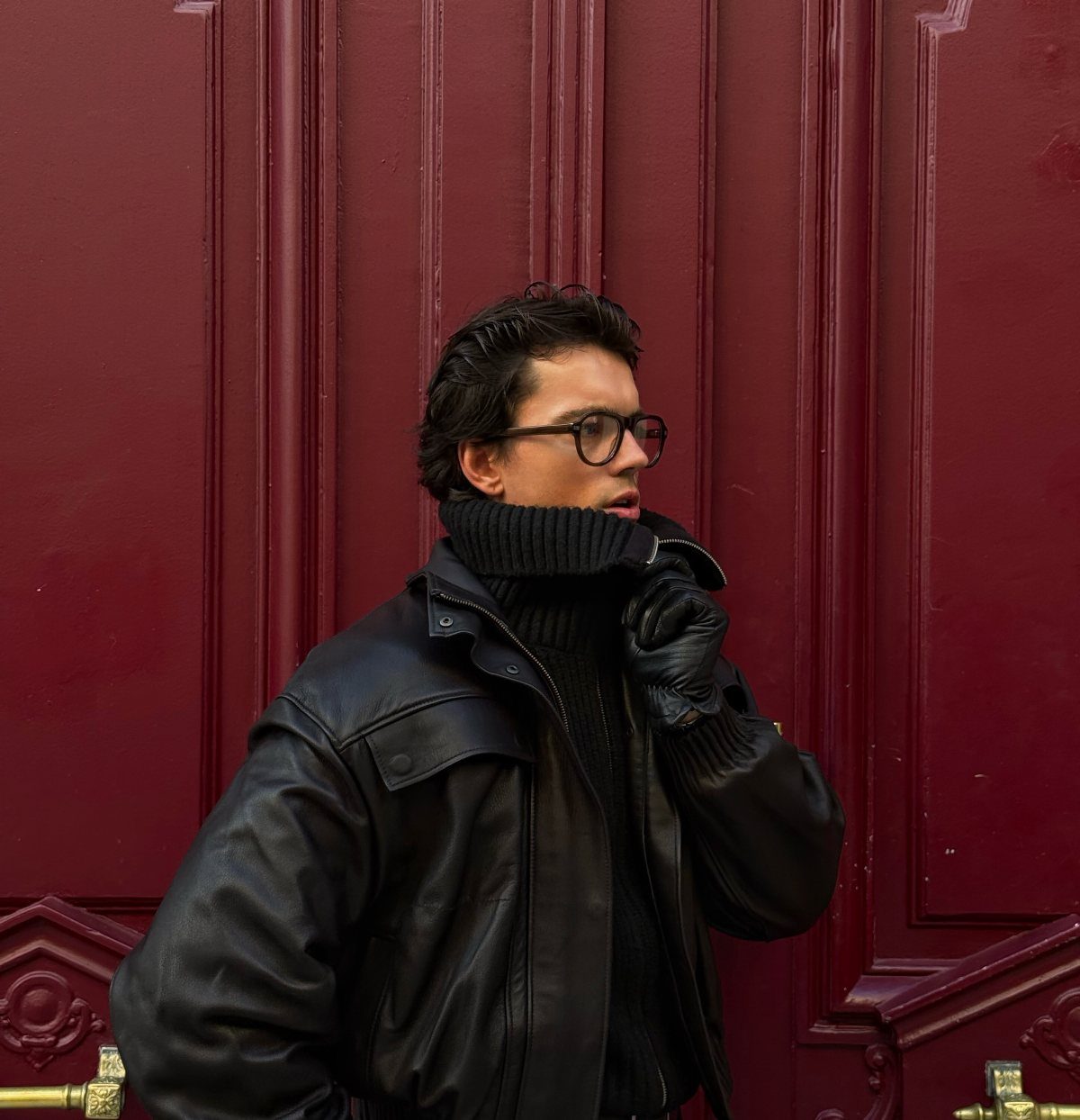 A man wearing a black turtleneck and leather jacket against a red door.