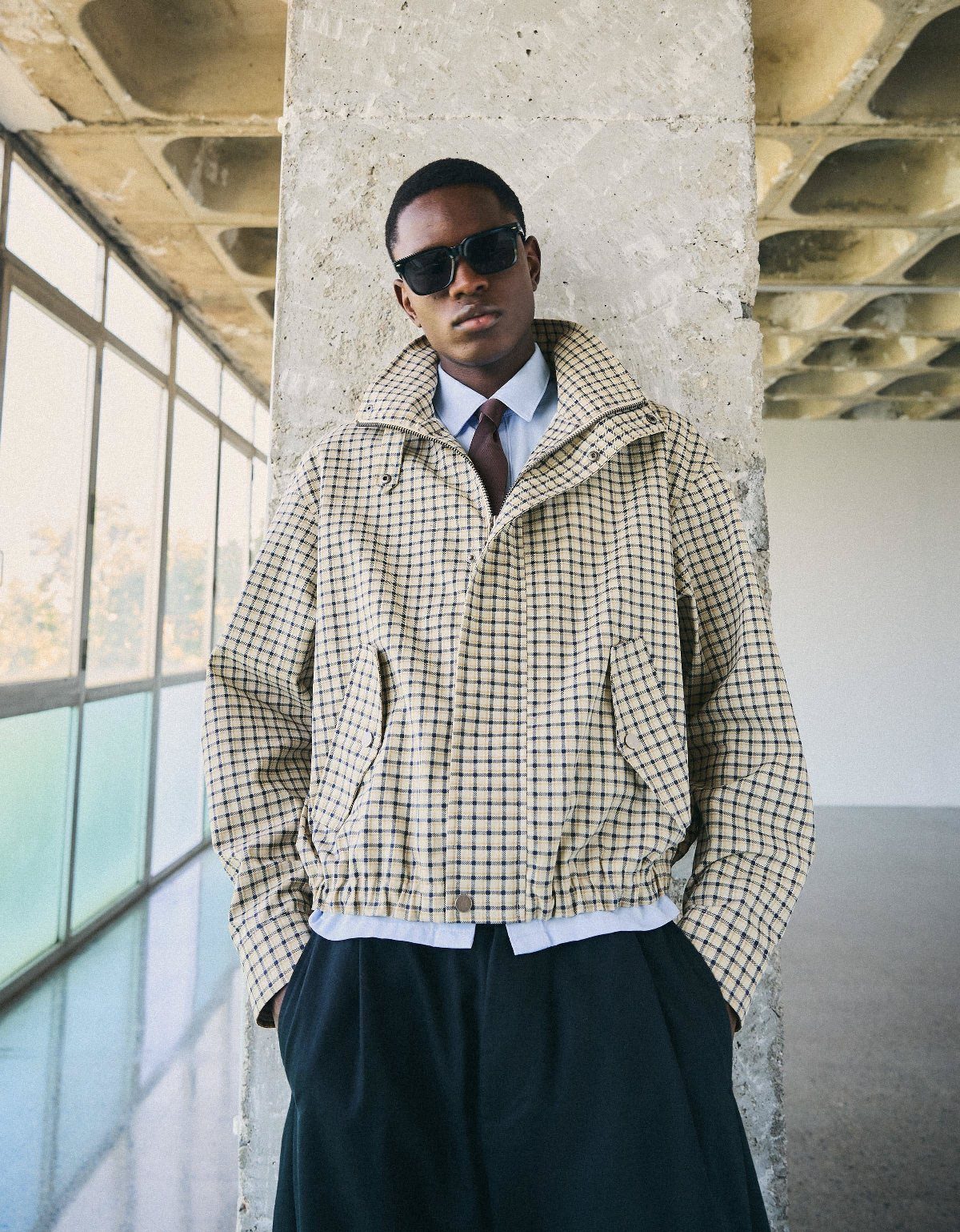 A man in a checkered patterned jacket and sunglasses leaning against a concrete pillar.