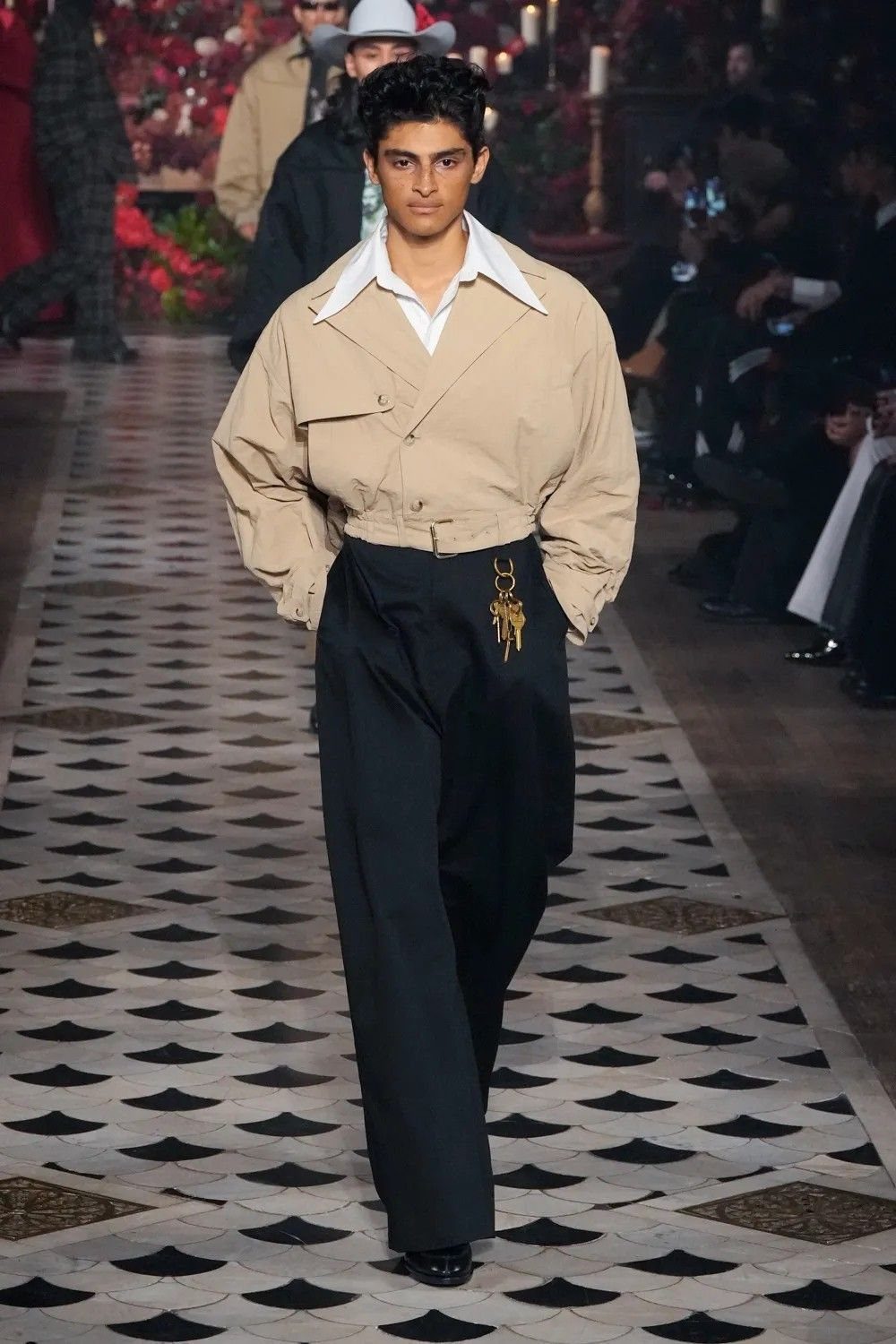 Male model walking a runway in a cropped beige shirt and wide-leg black trousers.