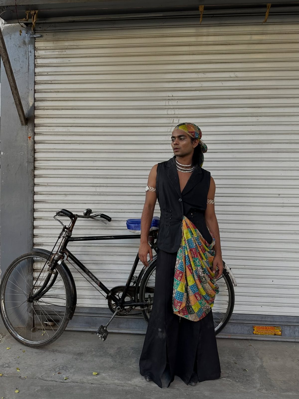 A person wearing a black vest, long trousers, and a colorful draped fabric stands by a bicycle.